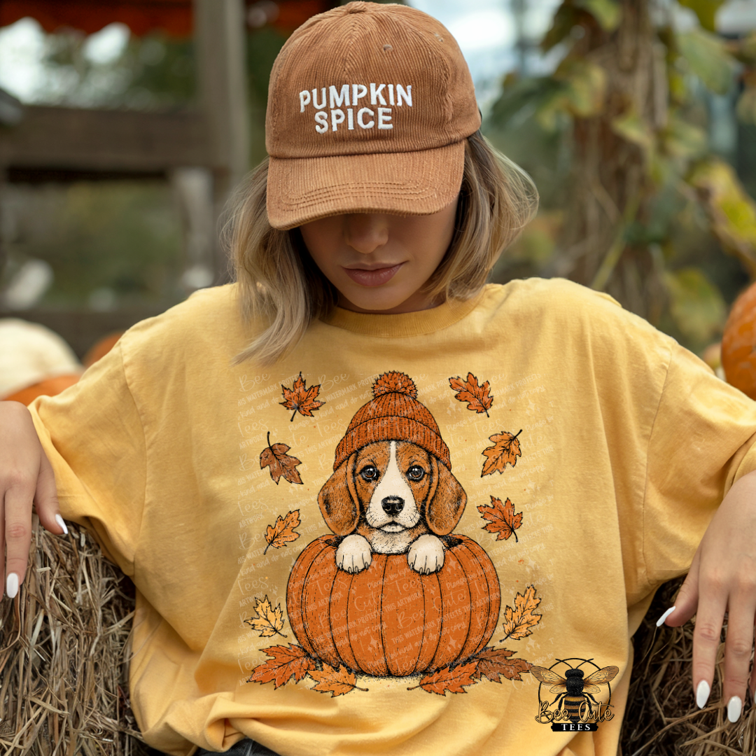 A woman wearing a tan 'Pumpkin Spice' baseball cap and a yellow graphic t-shirt featuring a puppy sitting on a pumpkin, posed on a hay bale at a pumpkin patch.