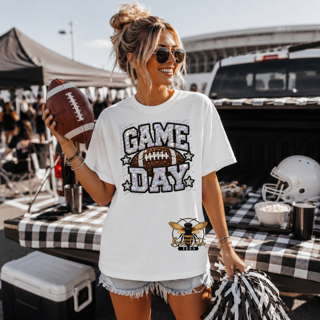 A woman wearing a white graphic t-shirt that says 'GAME DAY' with a football and bee logg, holding a football at an outdoor tailgate event.