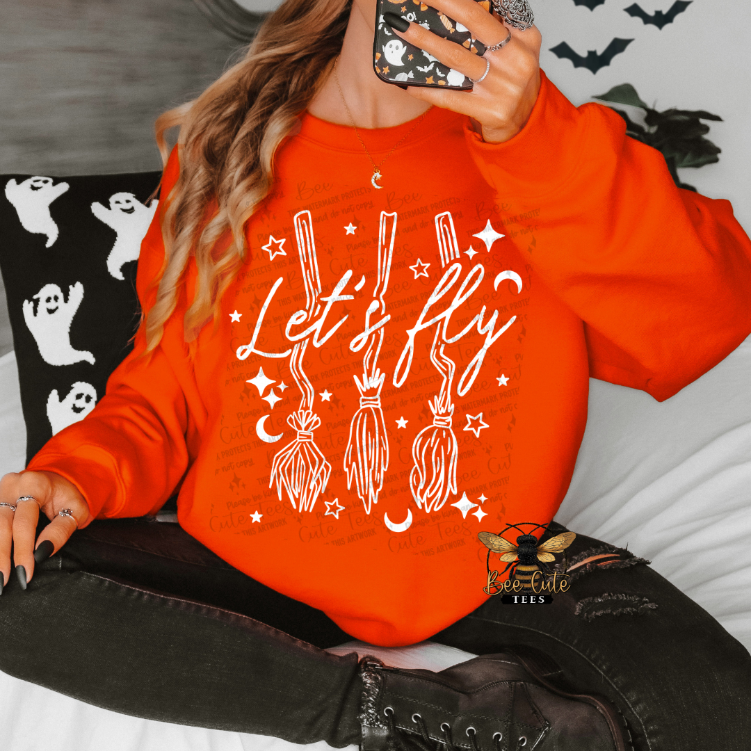 A woman wearing a bright orange crewneck sweatshirt with a white graphic that reads 'Let's fly' above three broomsticks, stars, and moons. She is sitting on a bed with a black pillow featuring white ghost illustrations.