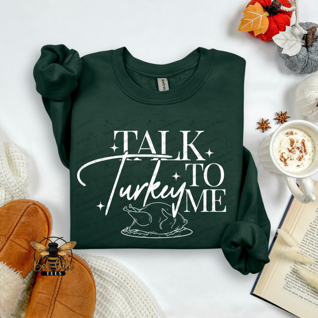 A dark forest green long-sleeve t-shirt with a white graphic reading 'TALK TO TURKEY' and a turkey illustration, styled in a flat lay with a mug of hot chocolate, an open book, knit socks, and autumn leaves.