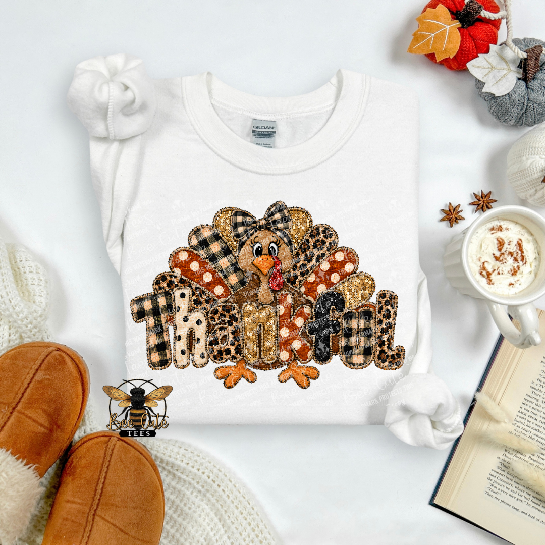 A folded white long-sleeve shirt with a large 'Thankful' turkey graphic in autumn patterns, styled in a cozy flat lay with a mug of coffee, knit socks, an open book, and autumn leaves.