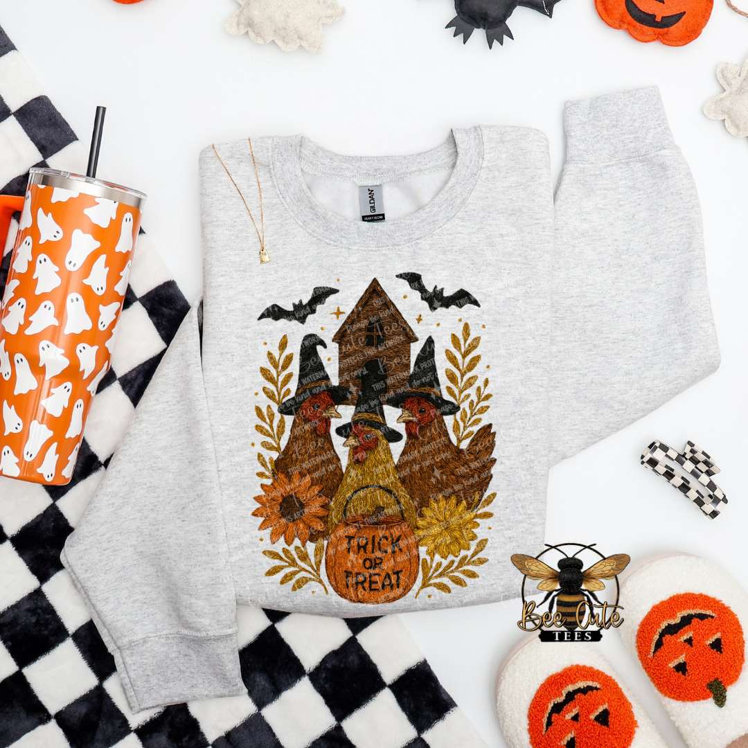 A light heather gray long-sleeve sweatshirt laid flat on a white background with Halloween-themed props. The sweatshirt features a large front graphic of chickens wearing witch hats under bats and a haunted house, with a pumpkin bucket that reads 'TRICK OR TREAT.