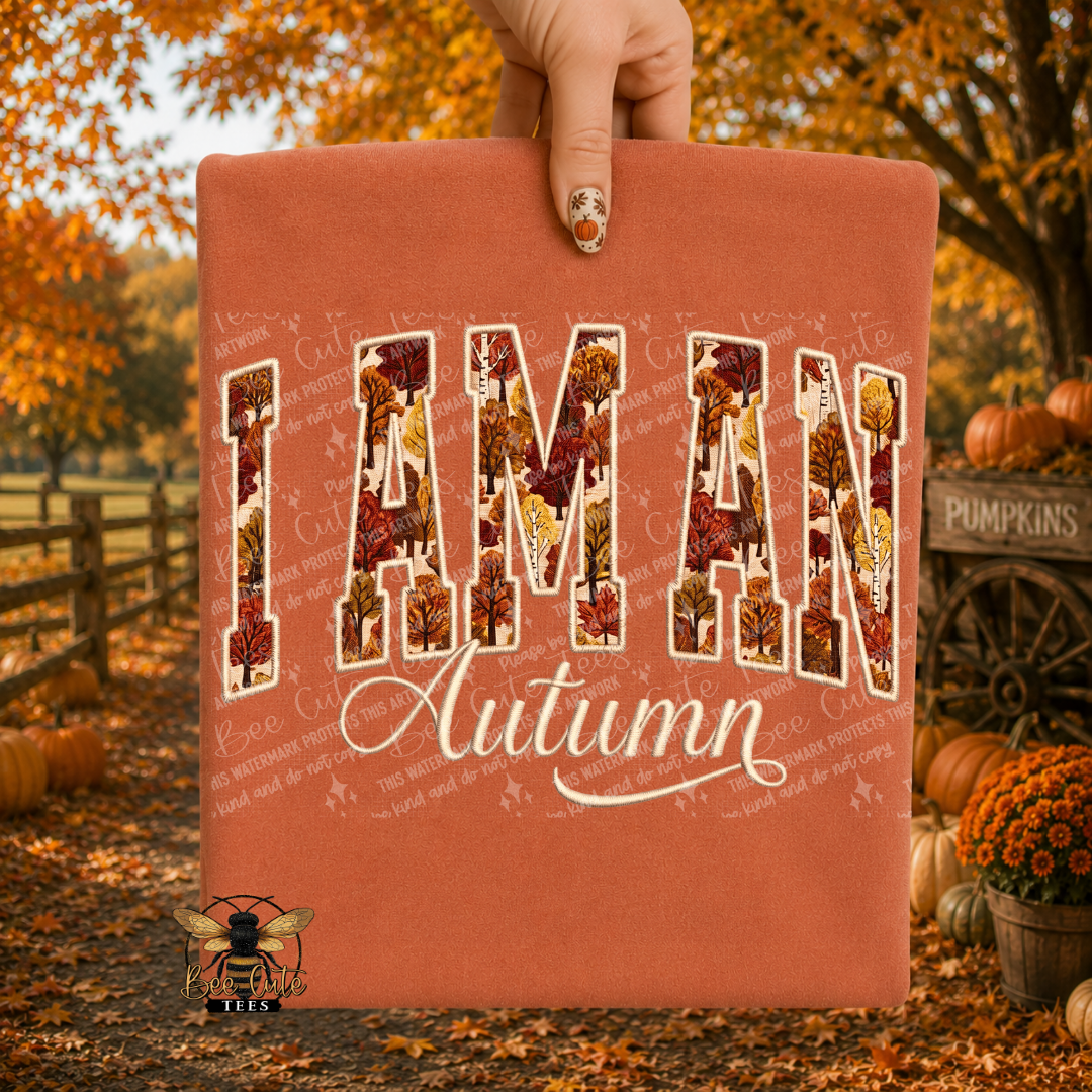 A folded terracotta-colored t-shirt held up by a hand, featuring a graphic that reads 'I AM AN Autumn' with 'I AM AN' filled with a fall leaf pattern. The background is a blurred autumn park scene with pumpkins and fall foliage.