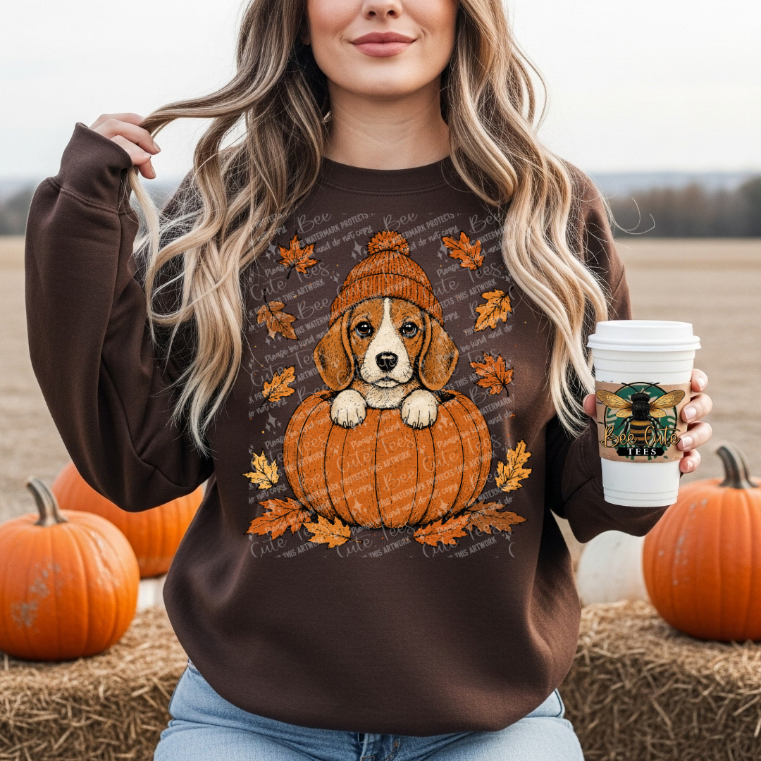 A woman wearing a brown crewneck sweatshirt with a graphic of a puppy sitting in an orange pumpkin, surrounded by autumn leaves. She is holding a coffee cup and standing outdoors on a hay bale with pumpkins in the background.