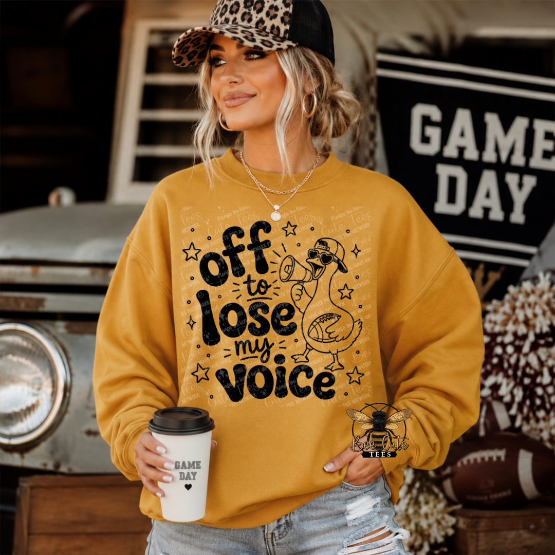 A woman wearing a mustard yellow sweatshirt with a black graphic that reads 'off to lose my voice' and a cartoon duck holding a football, standing in a rustic outdoor setting with a 'GAME DAY' sign and football props.