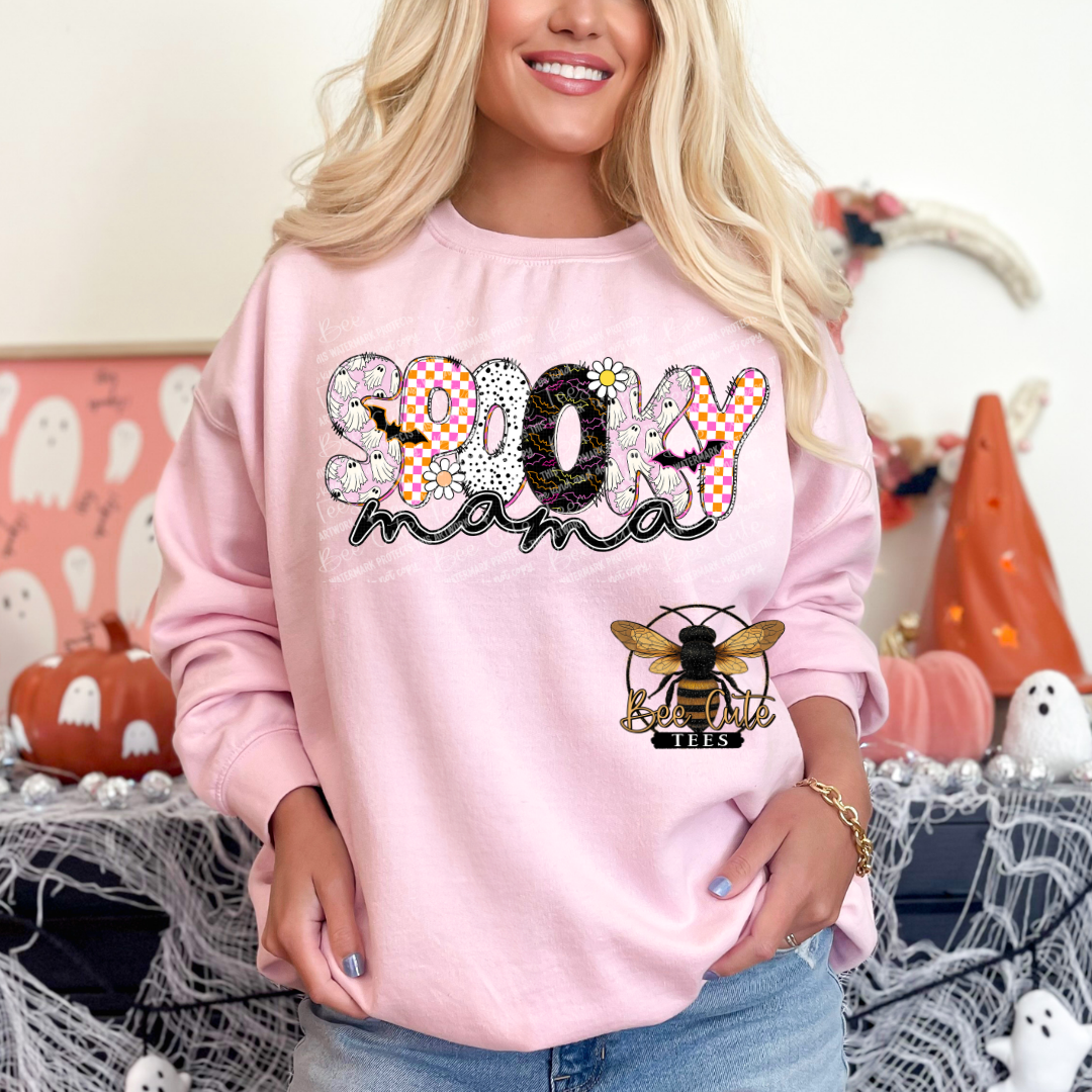 A woman wearing a light pink crewneck sweatshirt with a 'Spooky Mama' graphic and a bee design, posed in a Halloween-themed room with pumpkins and cobwebs.