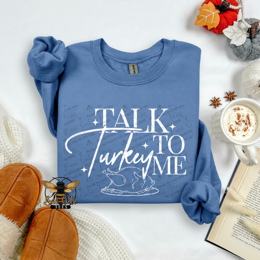 A folded blue long-sleeve sweatshirt with rolled cuffs, featuring a white graphic that reads 'TALK TO ME Turkey' with a turkey illustration. The sweatshirt is styled in a cozy flat lay with brown slippers, a mug of coffee, an open book, and autumn decorations on a white background.