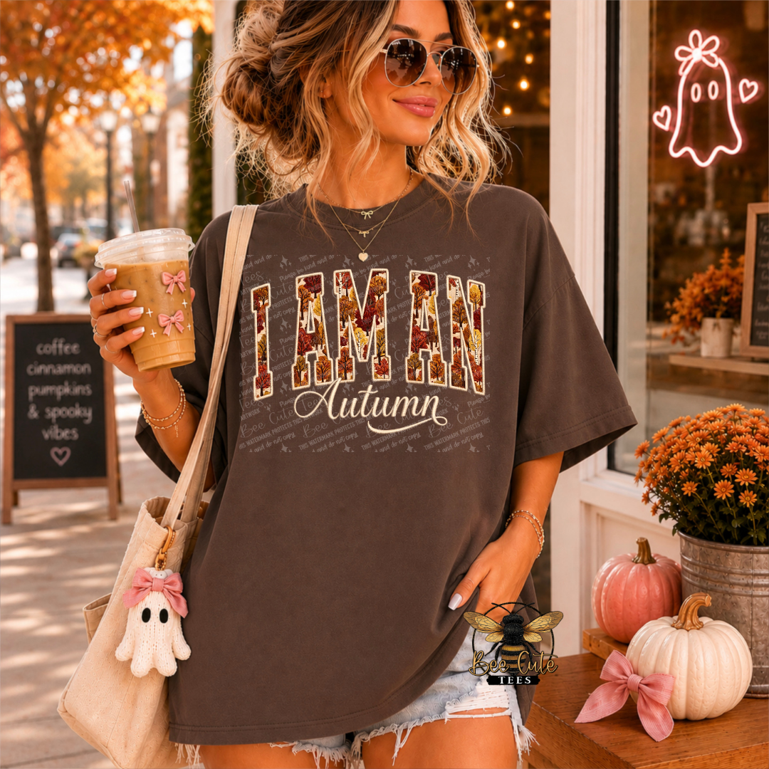 A woman wearing a dark brown graphic t-shirt with 'I AM AN Autumn' printed on the front in a distressed fall-themed font. She is holding a coffee cup and wearing a beige tote bag with a ghost charm, set against a cozy autumn storefront background with pumpkins and fall decorations.