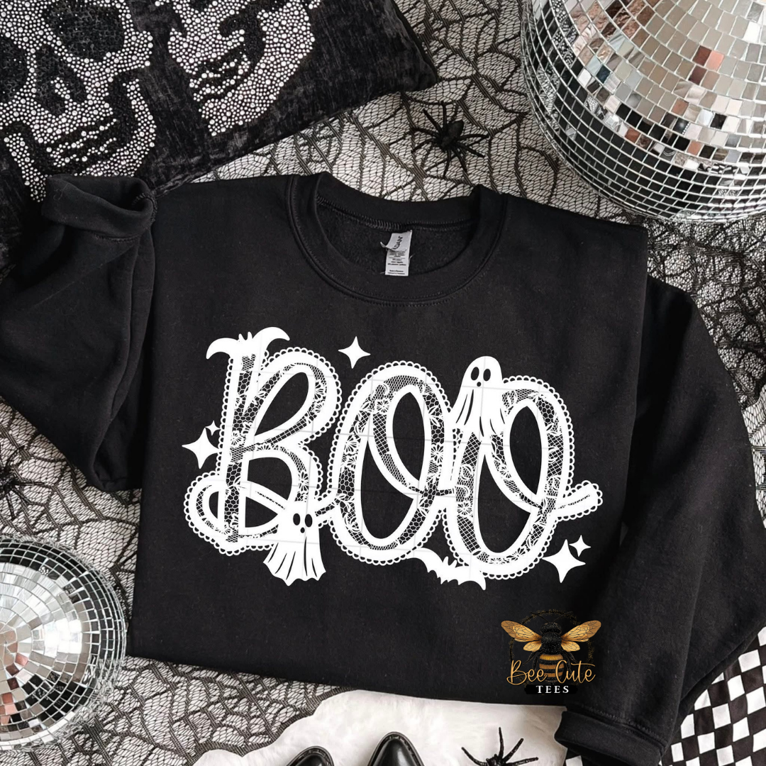 A black long-sleeve sweatshirt with a white 'BOO' graphic featuring ghosts, bats, and stars, styled in a Halloween-themed flat lay with spiderwebs, a disco ball, and a black rhinestone skull blanket.