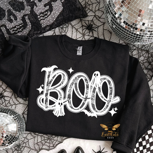 A black long-sleeve sweatshirt with a white 'BOO' graphic featuring ghosts, bats, and stars, styled in a Halloween-themed flat lay with spiderwebs, a disco ball, and a black rhinestone skull blanket.