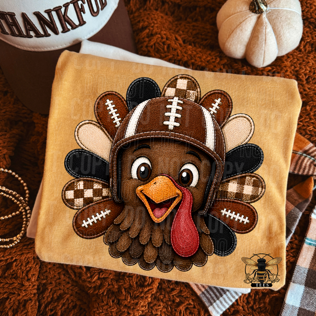 A square folded yellow T-shirt featuring a cartoon turkey wearing a football helmet, shown on a tan fabric background with autumn props including a pumpkin, plaid fabric, and a brown blanket.