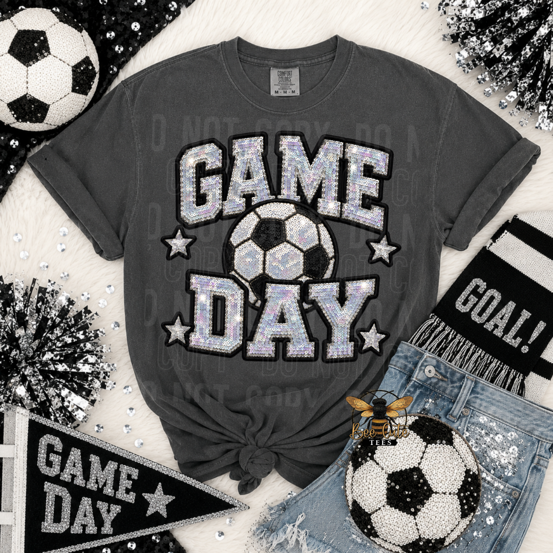 A dark gray graphic t-shirt featuring 'GAME DAY' in glittery silver letters with a soccer ball graphic in the center. The shirt is styled in a flat-lay arrangement with a black and white soccer ball, denim shorts, a black and white 'GOAL!' scarf, a 'GAME DAY' pennant, and silver pom-poms on a white faux fur background.