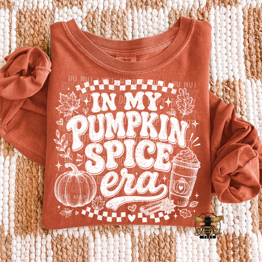 A rust-orange long-sleeve t-shirt with a white graphic reading 'IN MY PUMPKIN SPICE era' laid flat on a textured beige and white woven blanket. The graphic includes a pumpkin, cinnamon stick, hot cocoa cup, and autumn leaves.