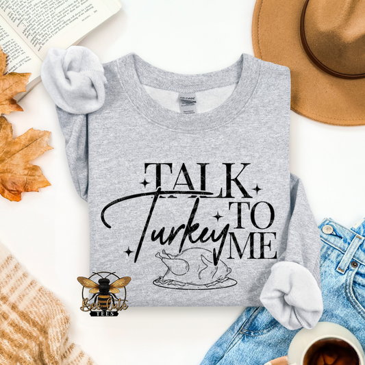 A folded heather gray graphic T-shirt is styled in a flat lay with an open book, autumn leaves, a tan hat, blue jeans, a cup of coffee, and a small bee logo. The shirt has black text reading “TALK Turkey TO ME” with a turkey graphic.