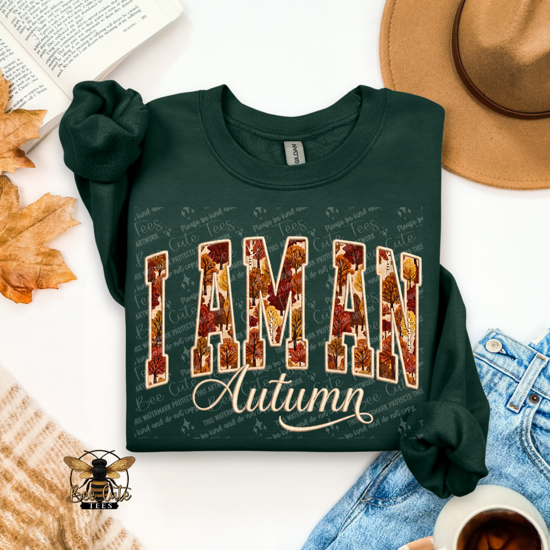 A dark green crewneck sweatshirt with a graphic reading 'I AM AN Autumn' in autumn-themed lettering, styled in a flat lay with a tan hat, blue jeans, a book, leaves, and a cup of coffee.