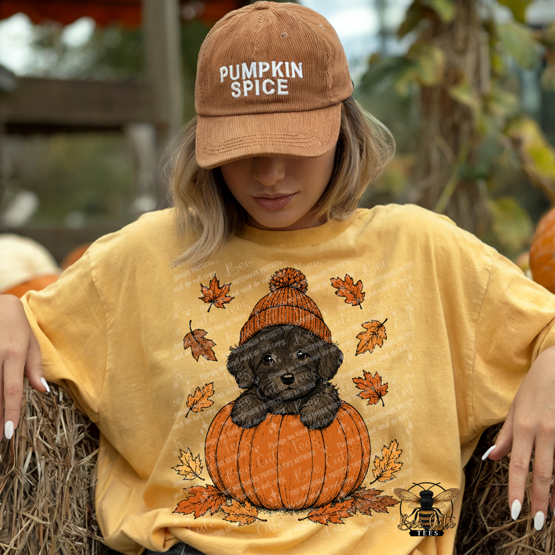 A woman wearing a tan 'Pumpkin Spice' baseball cap and a yellow graphic t-shirt featuring a black puppy sitting on a pumpkin, posed on a hay bale at a pumpkin patch.