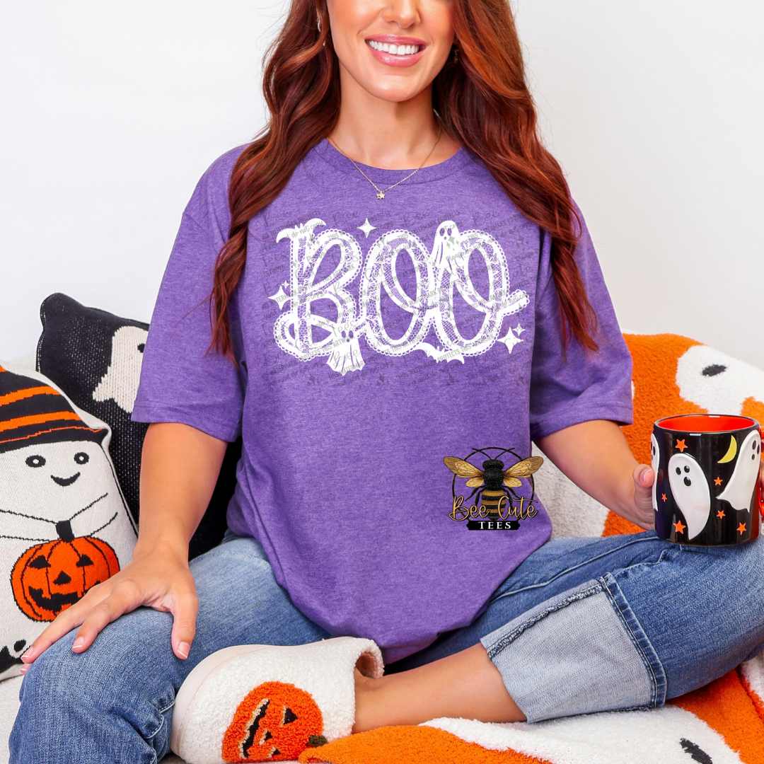 A woman wearing a purple heathered t-shirt with a white 'BOO' graphic and a bee logo that reads. She is sitting on a cozy orange and white blanket with Halloween-themed pillows.