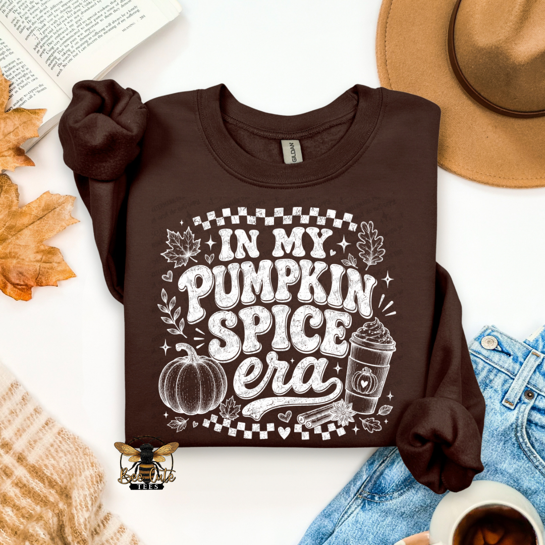 A dark brown long-sleeve graphic T-shirt laid flat on a white background with autumn and casual styling props, including a tan hat, blue jeans, a book page, leaves, a sweater, and a cup. The shirt has a large white graphic reading “IN MY PUMPKIN SPICE era” with a pumpkin, leaves, stars, and a coffee cup.