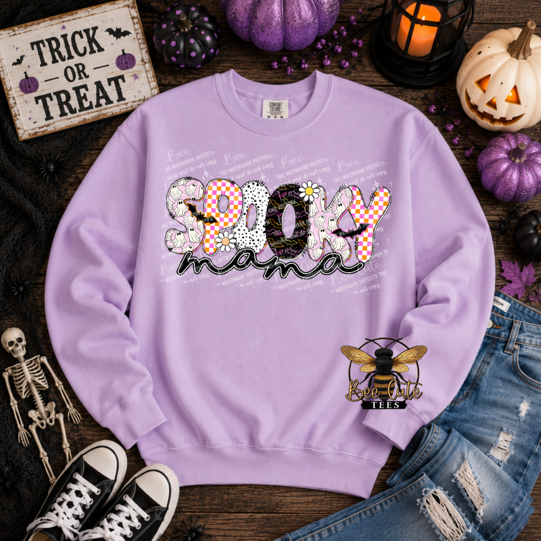 A lavender crewneck sweatshirt laid flat on a rustic wood surface, surrounded by Halloween props including pumpkins, a skeleton, jeans, sneakers, and a sign that reads 'Trick or Treat.' The sweatshirt has a large 'Spooky Mama' graphic across the chest with mixed patterned lettering and a bee logo on the lower right sleeve area.