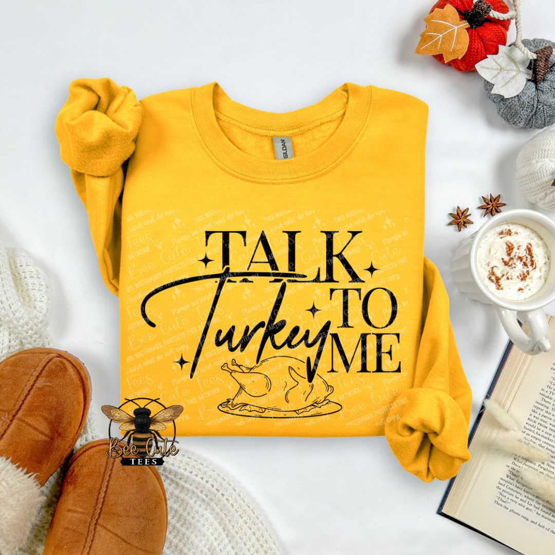A folded yellow sweatshirt with rolled sleeves on a white flat-lay background, featuring the black text 'TALK TO ME' and a turkey graphic. Surrounding props include brown suede shoes, a mug of coffee with whipped cream, an open book, and autumn leaves.
