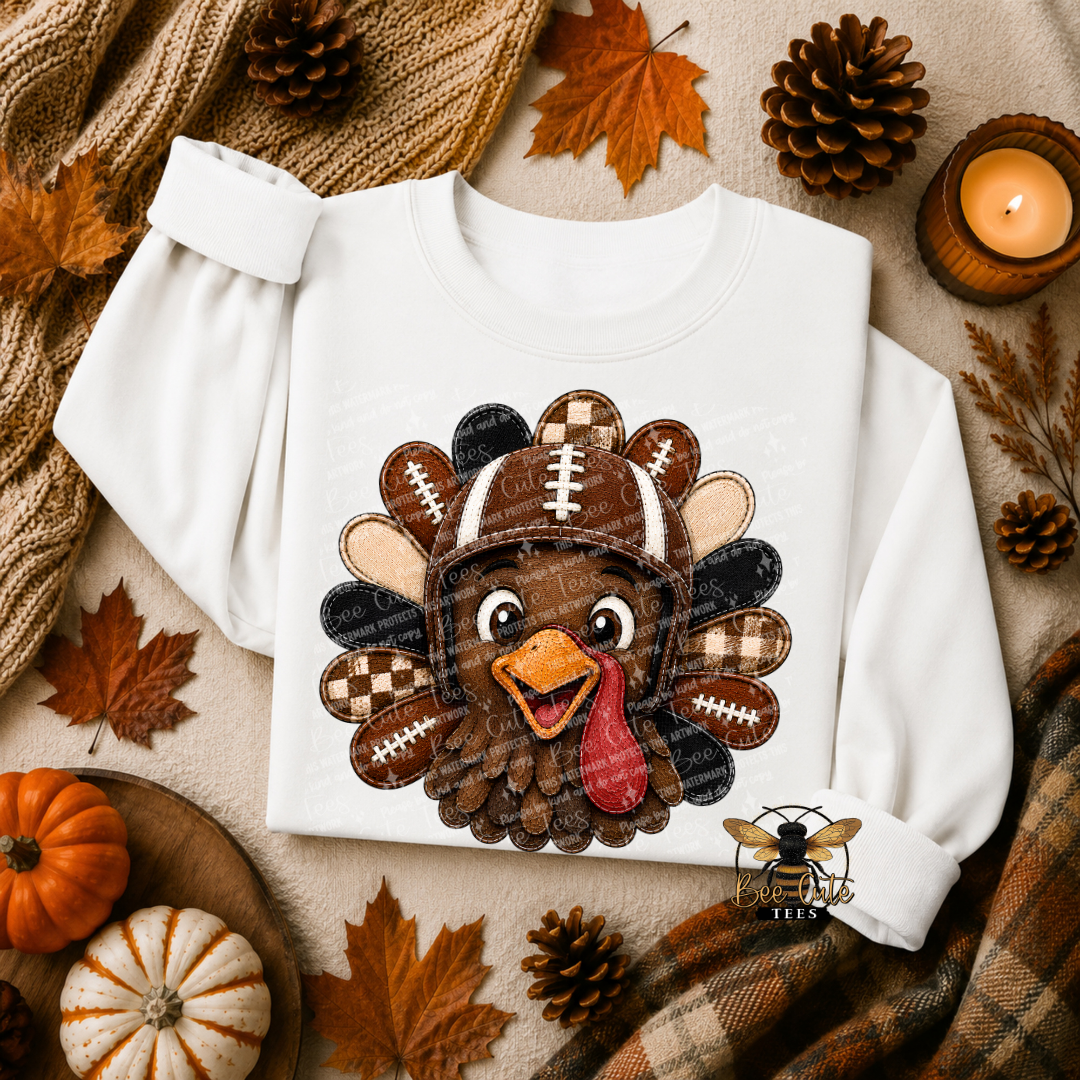 A white long-sleeve crewneck sweatshirt laid flat on a cozy autumn-themed background. The sweatshirt features a large applique-style graphic of a cartoon turkey wearing a football helmet.