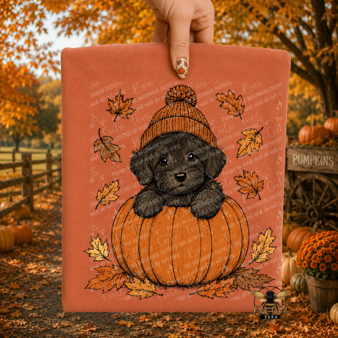 A hand holds a square fabric art print featuring a black puppy sitting in an orange pumpkin with autumn leaves and a knit hat, set against an outdoor fall yard scene with pumpkins and autumn trees.