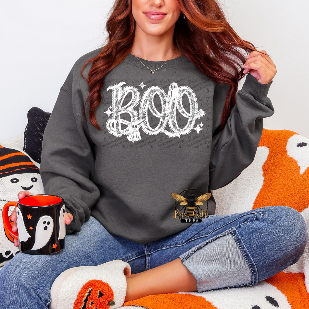 A woman sitting cross-legged on a white and orange patterned couch, wearing a dark gray crewneck sweatshirt with a white 'BOO' graphic printed on the front. She is holding a black and orange mug with a ghost design.