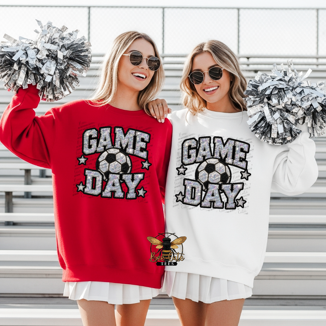 Two women standing on stadium bleachers wearing matching sweatshirts, one red and one white, both with a large 'GAME DAY' soccer graphic on the front. They are holding silver and white pom-poms.