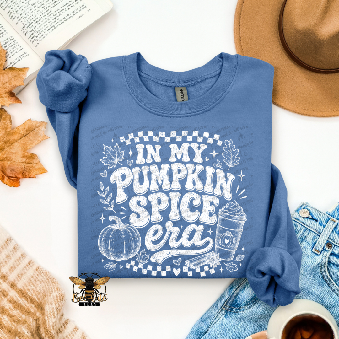 A folded blue long-sleeve graphic sweatshirt laid flat on a white surface with autumn and casual styling props, including a tan hat, denim shorts, a knit sweater, an open book, and a coffee cup. The shirt has a large white graphic reading “IN MY PUMPKIN SPICE era” with a pumpkin, leaves, stars, and checker details.
