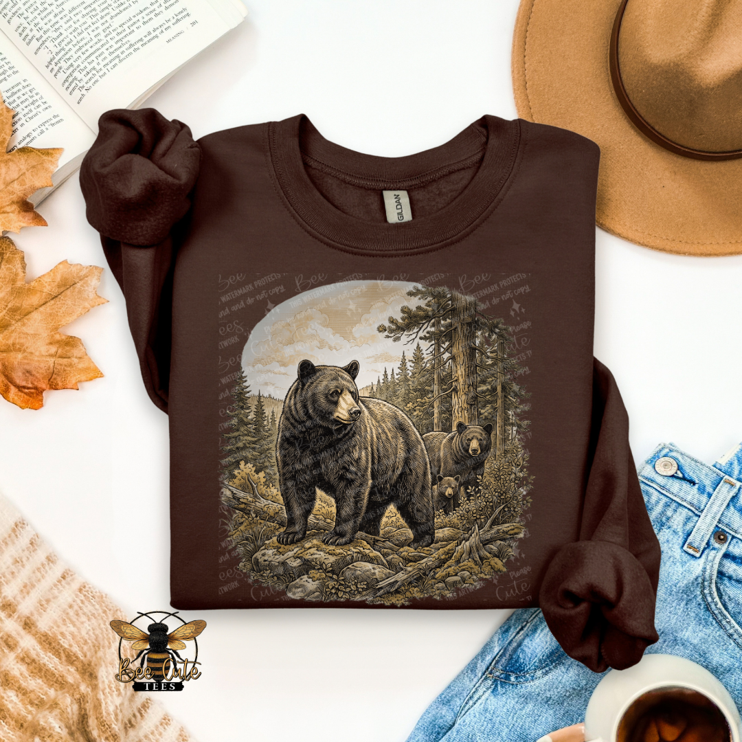 A dark brown crewneck sweatshirt with a detailed graphic of a black bear in a forest, styled in a flat-lay arrangement with autumn leaves, a tan hat, blue jeans, a cup of coffee, and an open book.