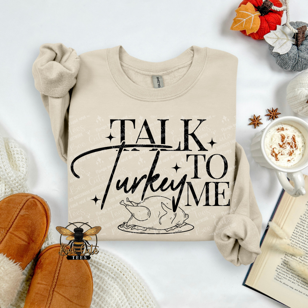 A beige crewneck sweatshirt with a black graphic reading 'TALK Turkey TO ME' and a turkey illustration, styled in a cozy flat lay with brown moccasin boots, a mug of hot cocoa, an open book, and autumn decorations on a white background.
