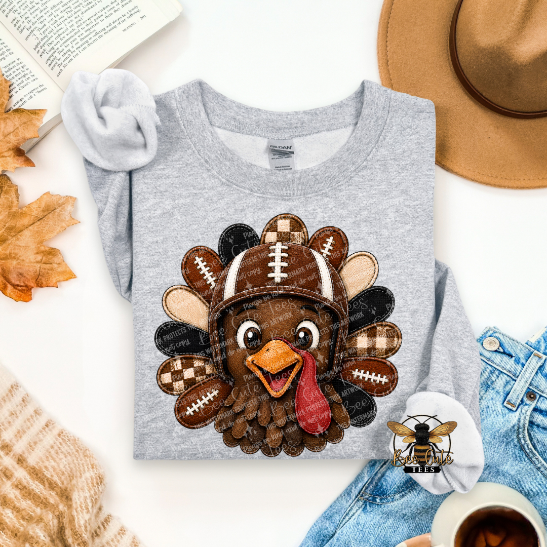 A folded heather gray long-sleeve shirt with a large applique turkey graphic wearing a football helmet, styled in a flat lay with autumn leaves, an open book, a tan hat, blue jeans, a cup of coffee, and a small bee graphic.