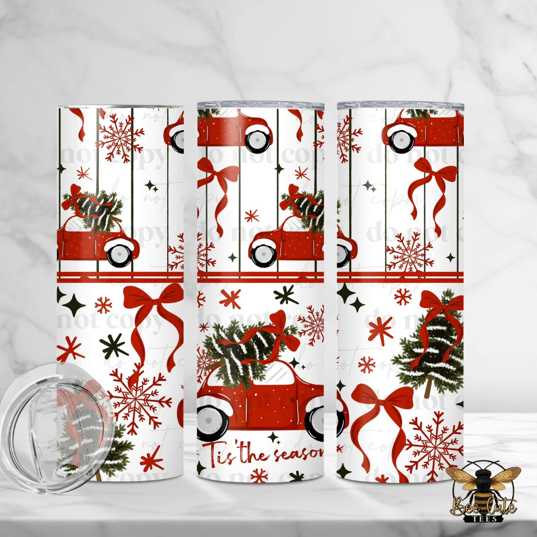 Three tall insulated tumblers with a red and white Christmas holiday print featuring vintage cars, pinecones, snowflakes, and red bows, shown on a light marble surface with a clear lid in the foreground.