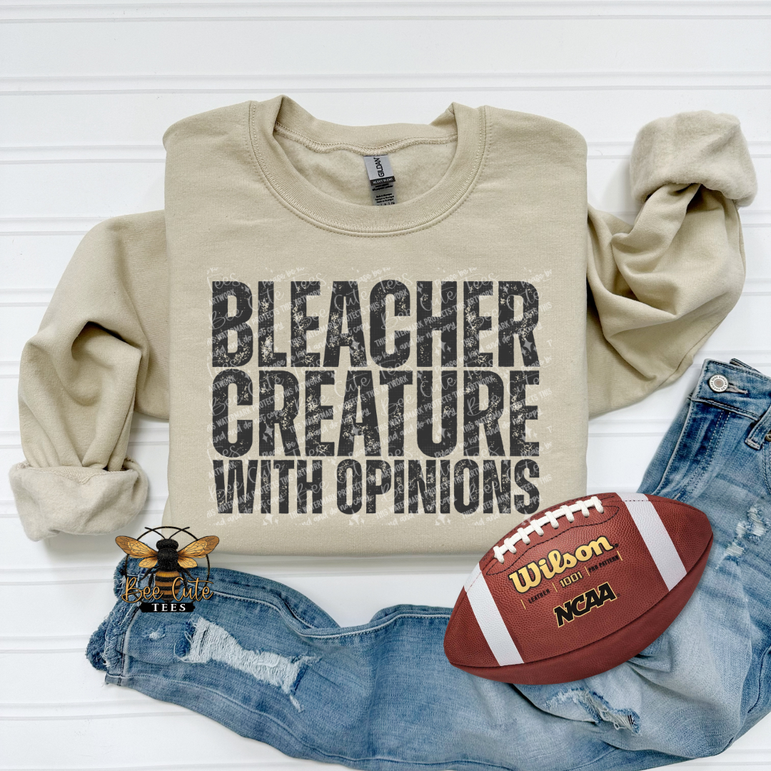 A beige crewneck sweatshirt with the text 'BLEACHER CREATURE WITH OPINIONS' in distressed black lettering, styled in a flat lay with blue jeans and a  football on a white wood background.
