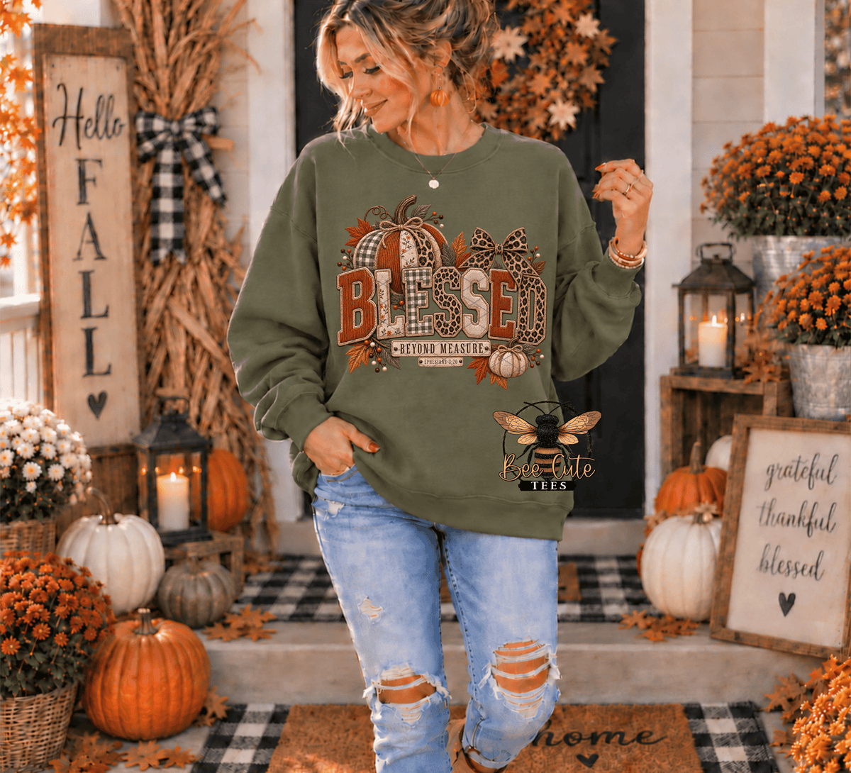 A woman wearing an olive green long-sleeve sweatshirt with a 'Blessed' autumn graphic, paired with distressed light blue jeans, standing in a decorated fall-themed front yard.