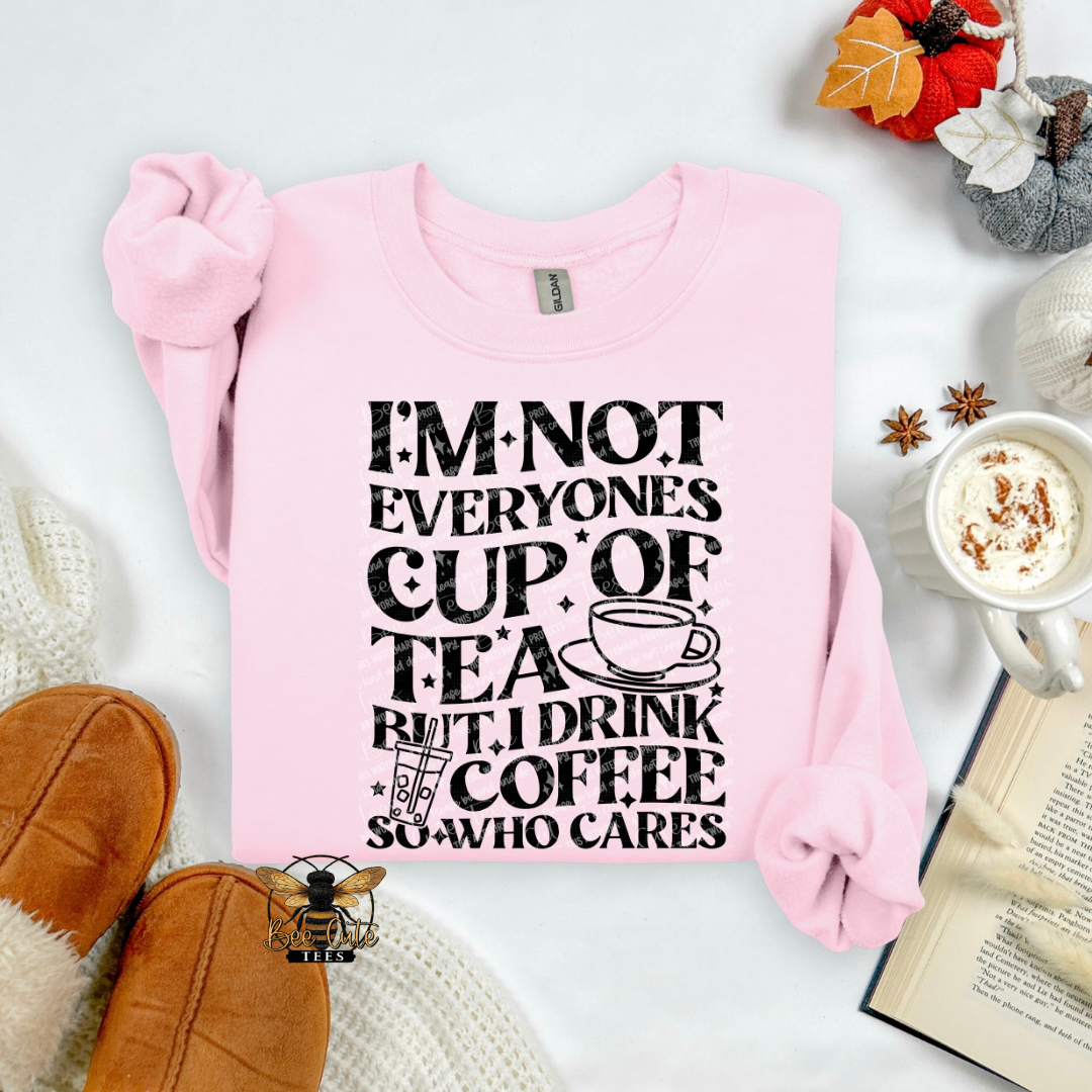 A light pink crewneck sweatshirt with a black graphic text print reading 'I'M NOT EVERYONE'S CUP OF TEA BUT I DRINK COFFEE SO WHO CARES' with a coffee cup illustration. The sweatshirt is styled in a cozy flat-lay arrangement with brown slippers, a mug of coffee, an open book, and autumn leaves.