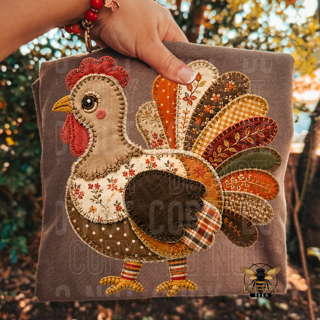A hand holds a square brown folded t-shirt featuring a colorful patchwork applique turkey. The background shows a blurred outdoor garden setting with trees and sunlight.