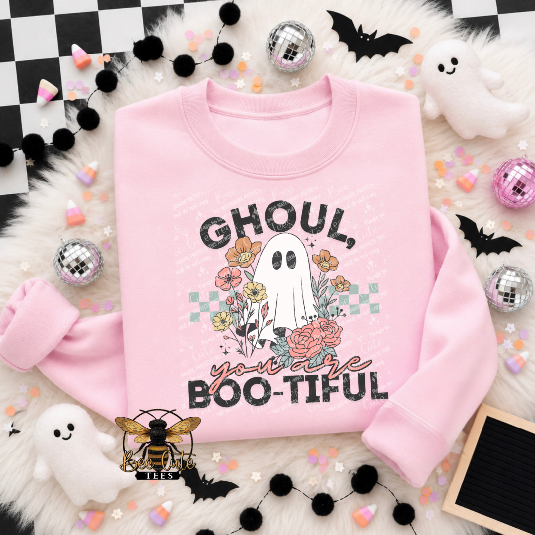 A pink crewneck sweatshirt laid flat on a white faux-fur surface with Halloween-themed props, including black pom-pom garlands, silver disco balls, candy corn, ghost plushies, bat cutouts, and a checkered corner. The sweatshirt has a ghost and floral graphic with the text 'GHOUL you are BOO-TIFUL'.