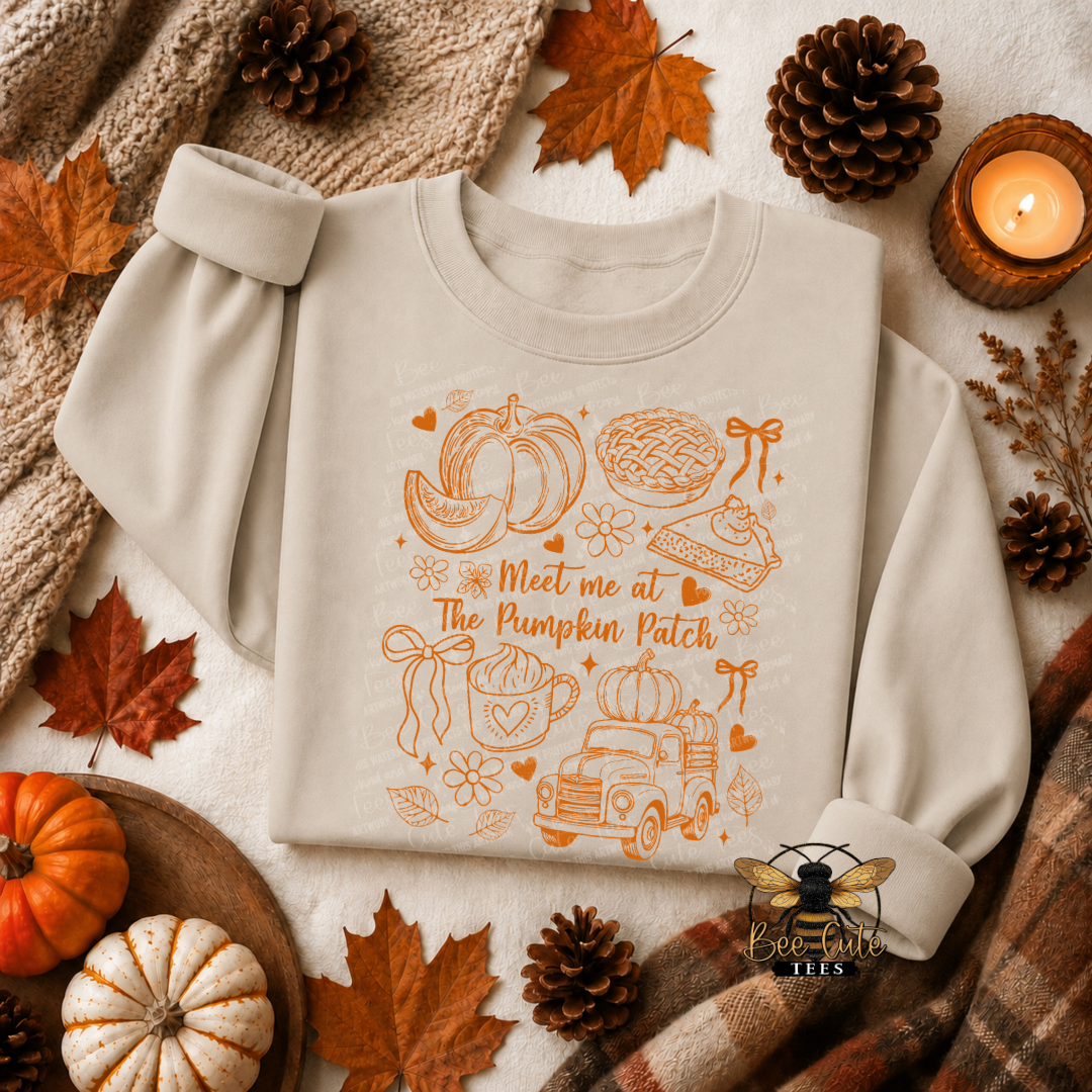 A cream-colored long-sleeve sweatshirt with an orange autumn-themed graphic reading 'Meet me at The Pumpkin Patch' and featuring illustrations of pumpkins, a truck, a pie, and a mug. The sweatshirt is styled in a cozy flat-lay arrangement with fall leaves, pinecones, a knitted blanket, a small pumpkin, and a lit candle.