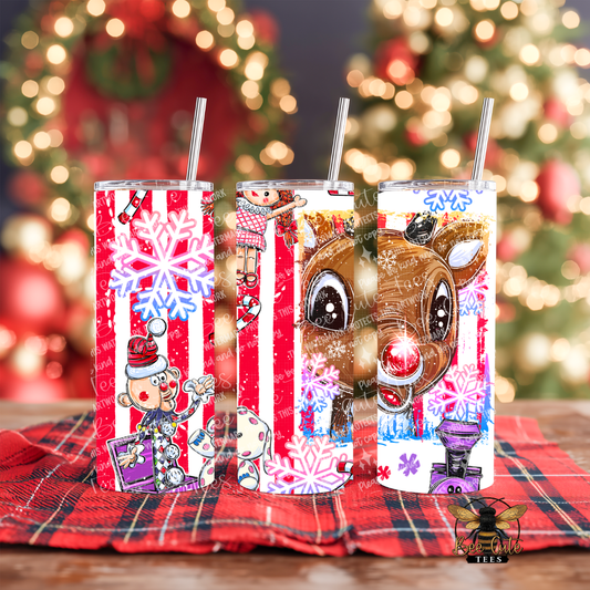 Three festive holiday-themed tumbler cups with straws, decorated with red-and-white stripes, snowflakes, cartoon characters, and a reindeer face, displayed on a plaid cloth in front of a blurred Christmas tree background.