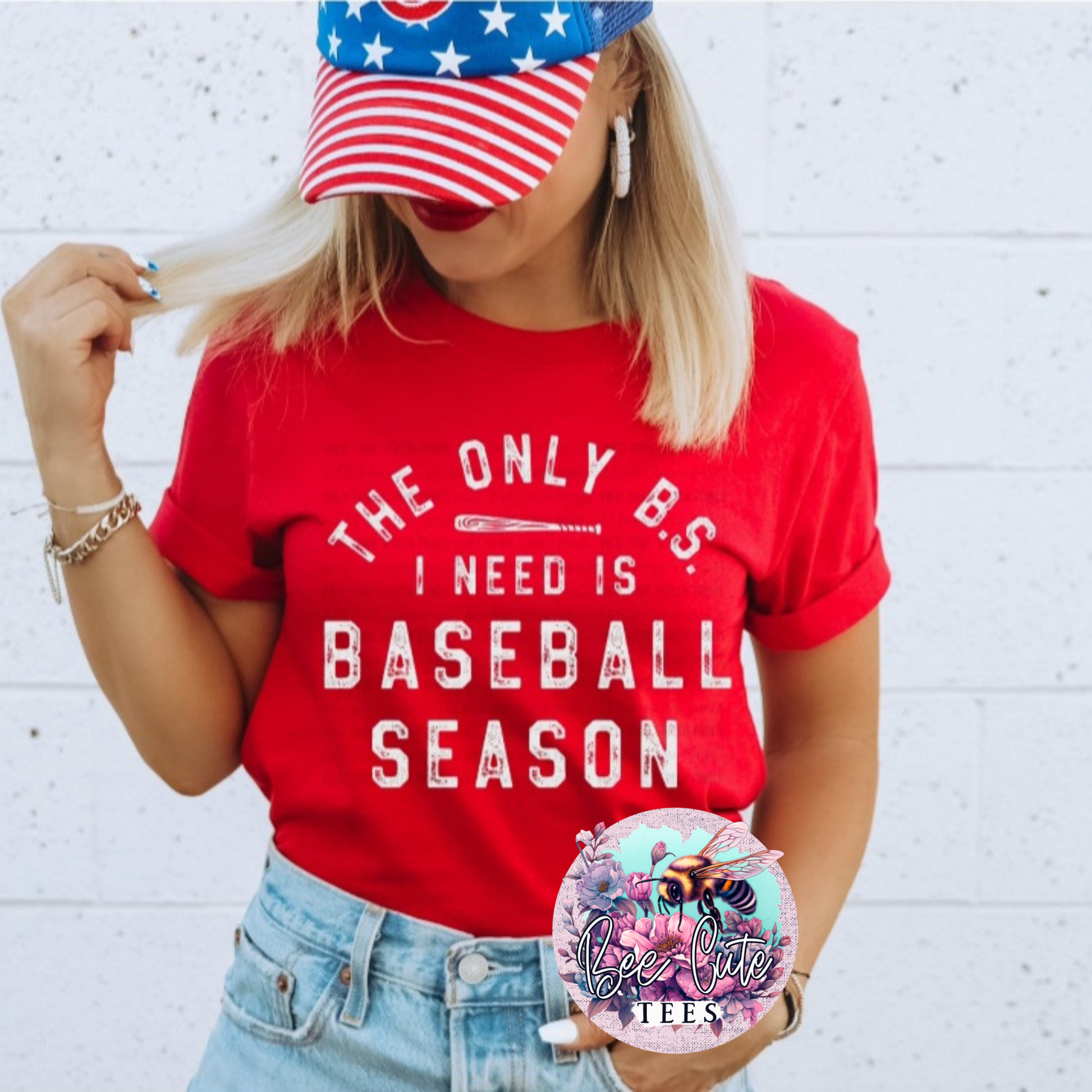 Person wearing a red t-shirt with 'The Only BS I Need is Baseball Season' text, an American flag hat, and denim shorts against a white brick wall.