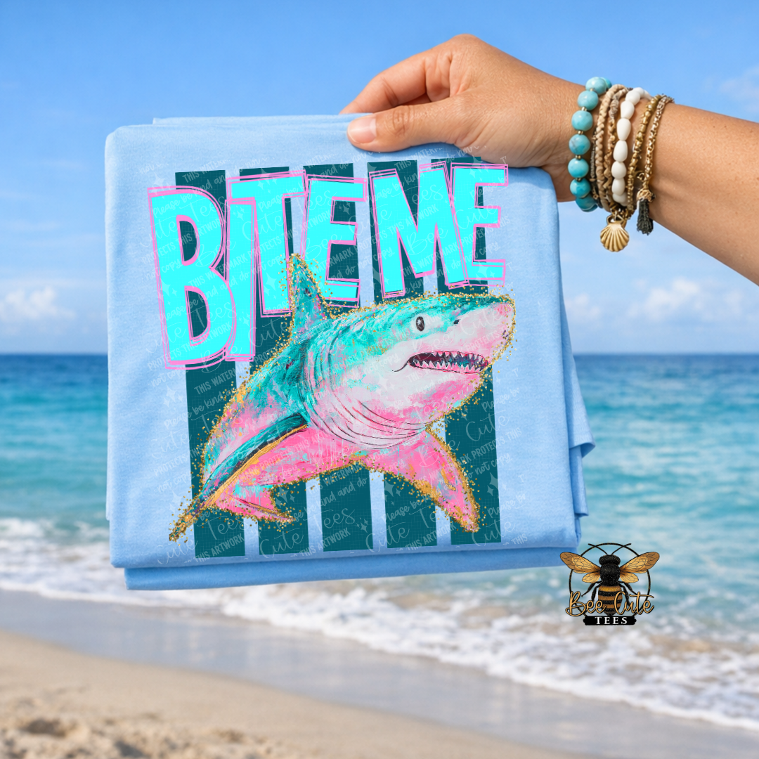 Light Blue t-shirt with shark design and 'BITE ME' text held by a hand on a beach.