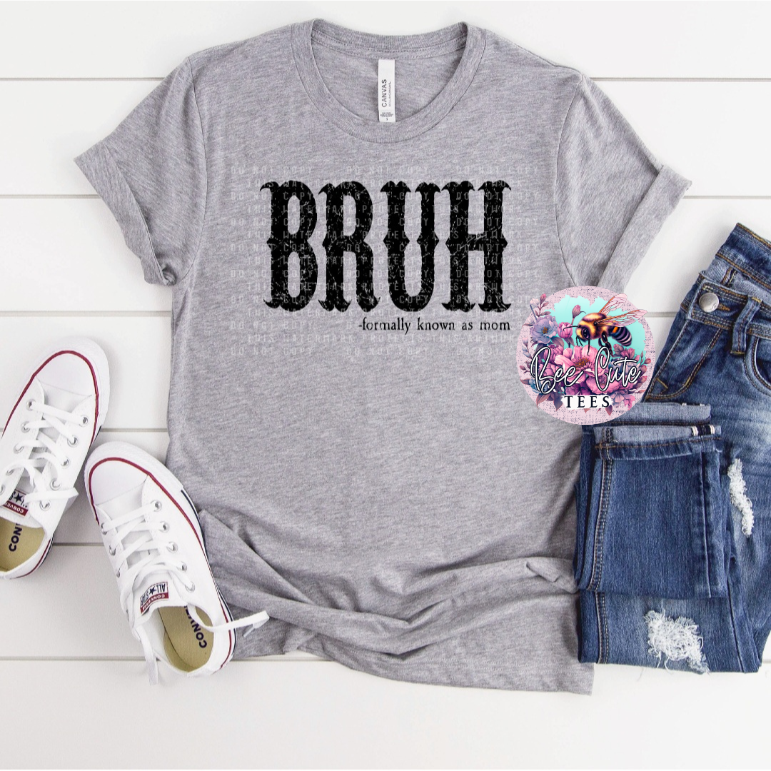 Gray t-shirt with 'BRUH formally known as mom' text, white sneakers, and jeans on a light wooden surface.