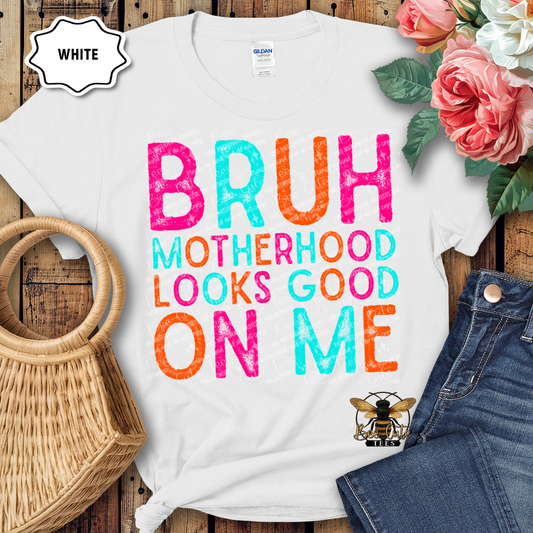 White t-shirt with colorful text 'BRUH MOTHERHOOD LOOKS GOOD ON ME' on a wooden surface with flowers and jeans.