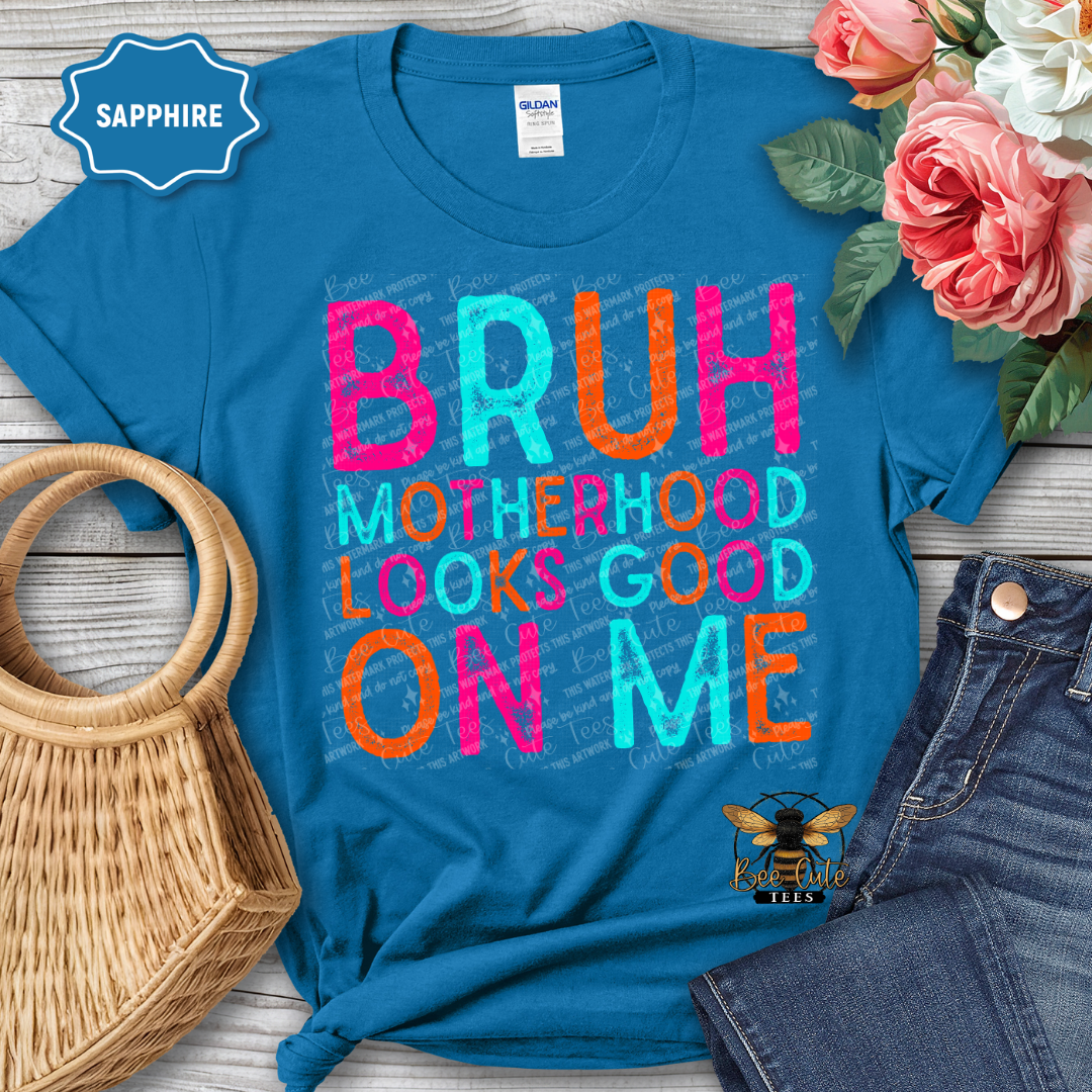Blue t-shirt with colorful text 'BRUH MOTHERHOOD LOOKS GOOD ON ME' on a wooden surface with flowers and denim jeans.