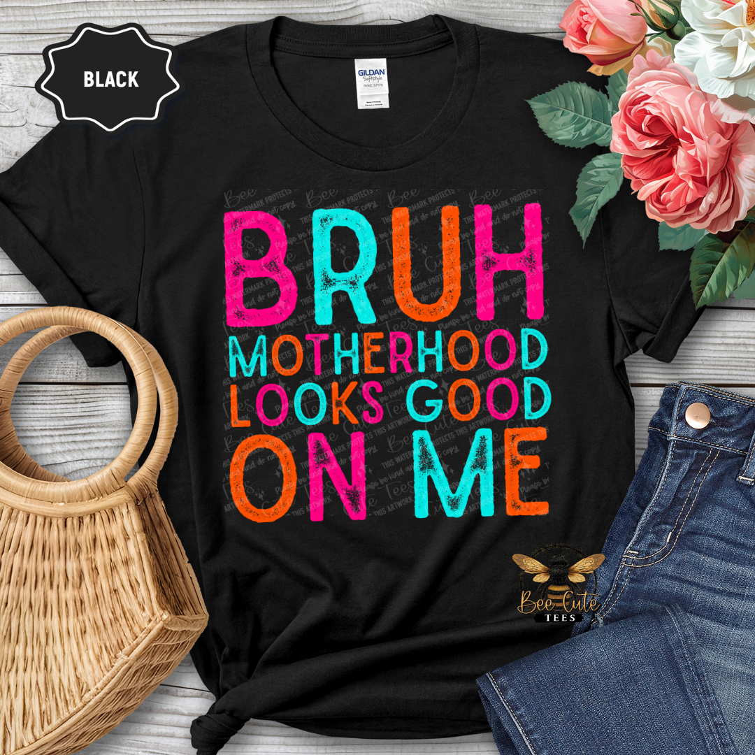 Black t-shirt with colorful text 'BRUH MOTHERHOOD LOOKS GOOD ON ME' on a wooden surface with flowers and jeans.