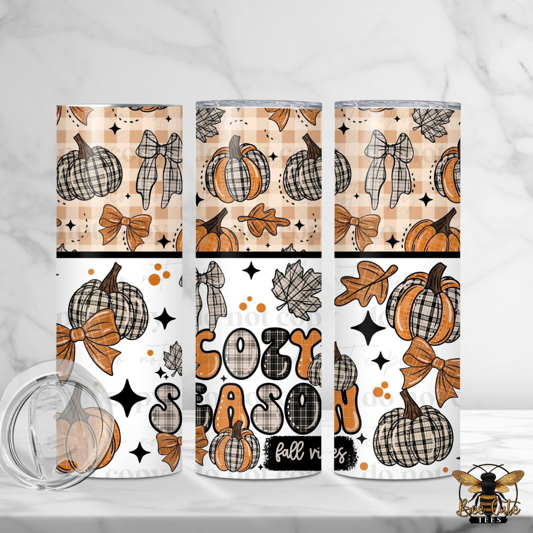 Three insulated tumblers with a fall-themed pattern of plaid pumpkins, bows, leaves, stars, and the words 'Cozy SEASON' on a white background. A clear lid is shown in front of the tumblers on a light marble surface.