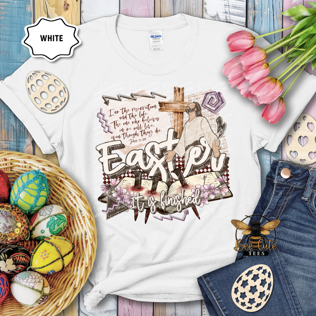 White t-shirt with Easter design featuring a cross and text, surrounded by Easter decorations on a wooden surface.