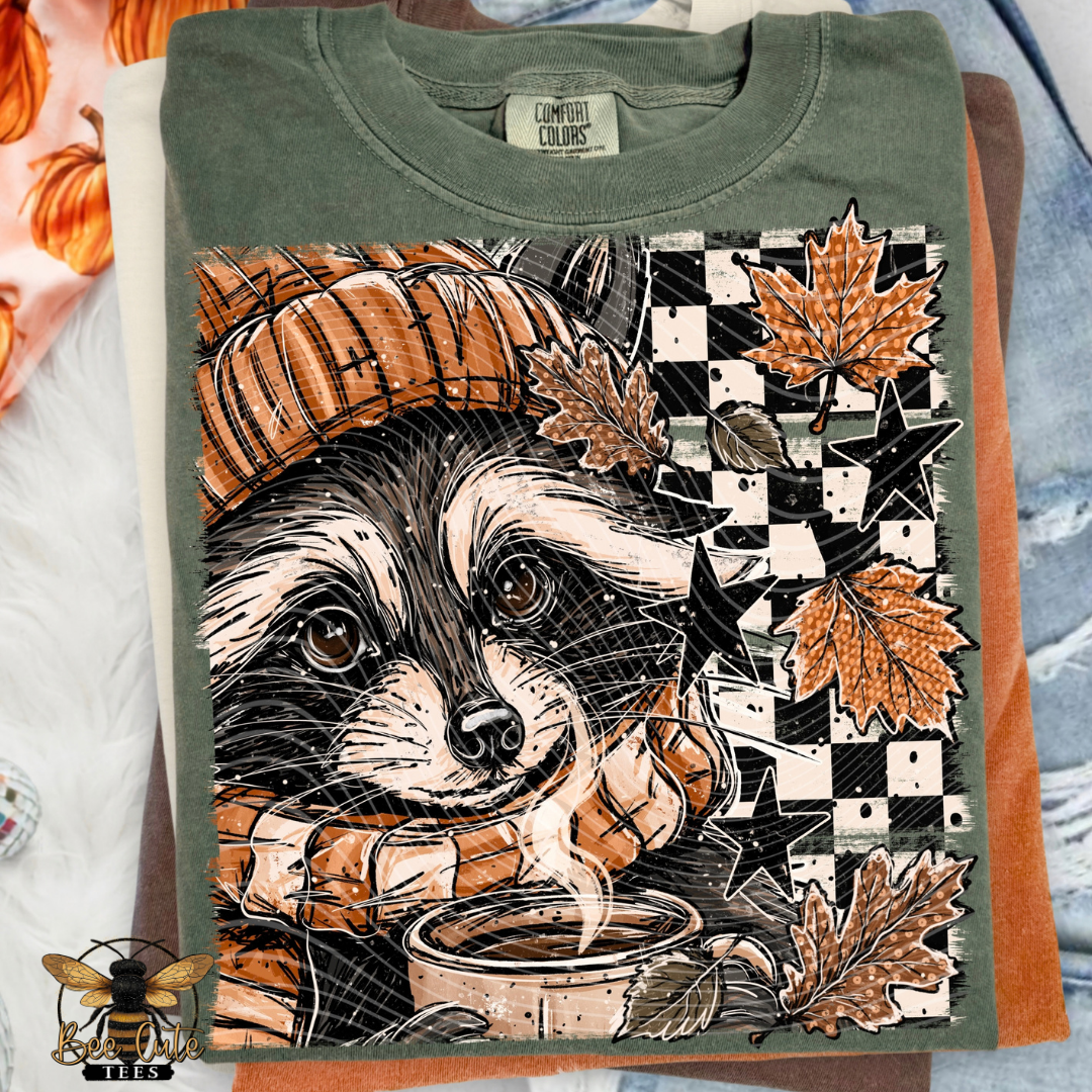 An olive green t-shirt featuring a detailed graphic print of a raccoon wearing an orange beanie and holding a mug, surrounded by autumn leaves, stars, and a checkerboard pattern. The shirt is folded and layered over other t-shirts, with a brand label visible inside the collar.