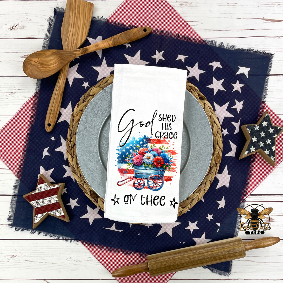 White kitchen towel displayed on a patriotic table setting, featuring a red, white, and blue wagon filled with flowers and the phrase “God Shed His Grace on Thee,” surrounded by star-patterned linens, wooden utensils, and rustic decor.
